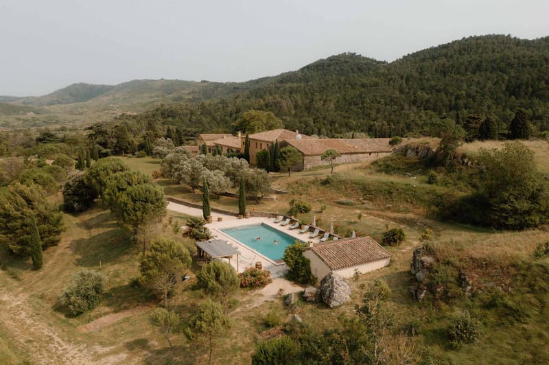 An aerial wide shot showcasing a large Provençal mas-style property with terracotta-tiled roofing and warm ochre stone walls, set within an expansive rural landscape of rolling hills and forested ridgelines. The grounds feature a rectangular swimming pool with turquoise water — one or two people are visible swimming — surrounded by light blue sun loungers and white parasols arranged along a stone terrace. Tall cypress trees line the approach to the main house, and the property includes several outbuildings, a covered pergola structure adjacent to the pool, and terracotta pot plantings. The overall palette of the grounds is dry gold and muted green, consistent with a southern French summer setting. Potential venue feature image.