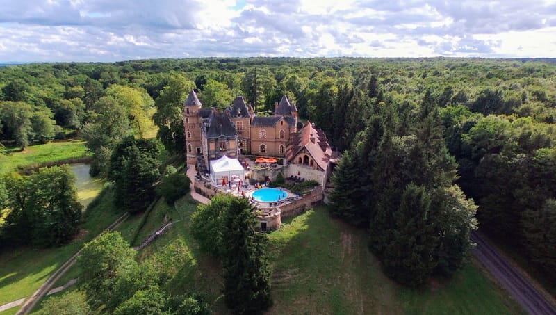 Aerial drone shot of a French château set within an extensive forested landscape, captured during what appears to be a wedding event. The château features multiple pointed turrets, warm sandstone and terracotta-tiled architecture, and a walled courtyard. A large white reception marquee tent has been erected in the courtyard beside the château, accompanied by orange parasols and a round above-ground pool with bright blue water. A small number of guests are visible around the pool and courtyard area. A pond or small lake is visible to the left of the property, and a road borders the right edge of the grounds. Potential venue feature image.