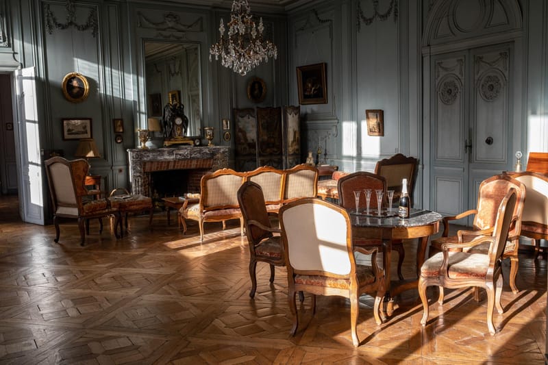 Chateau salon with blue-grey boiserie panelling, crystal chandelier, marble fireplace, and champagne service