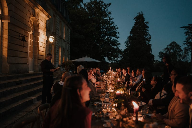 Candlelit banquet along chateau facade at dusk speaker at head of table bride visible mid-table among thirty guests