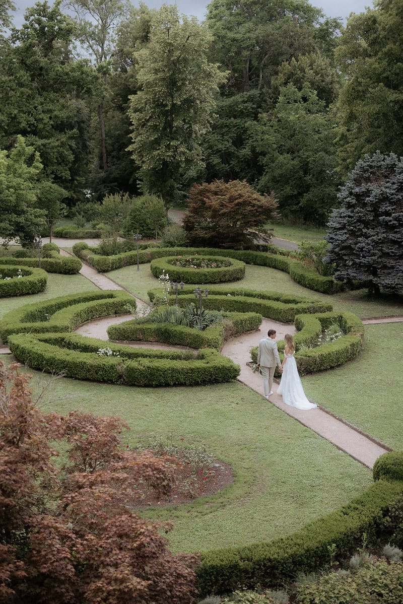 Aerial view of bride and groom walking hand-in-hand through a formal French parterre garden with curved boxwood hedges