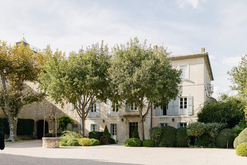 Wide exterior shot of a French country estate featuring a three-story rendered manor house with pale cream walls, soft grey-blue shutters, and terracotta roof tiles. The facade is flanked by mature trees and neatly trimmed boxwood hedges, with a gravel forecourt and a stone well visible to the left. An adjacent stone outbuilding with arched doorways is partially visible on the left side of the frame. No people are present in the image. Potential venue feature image.