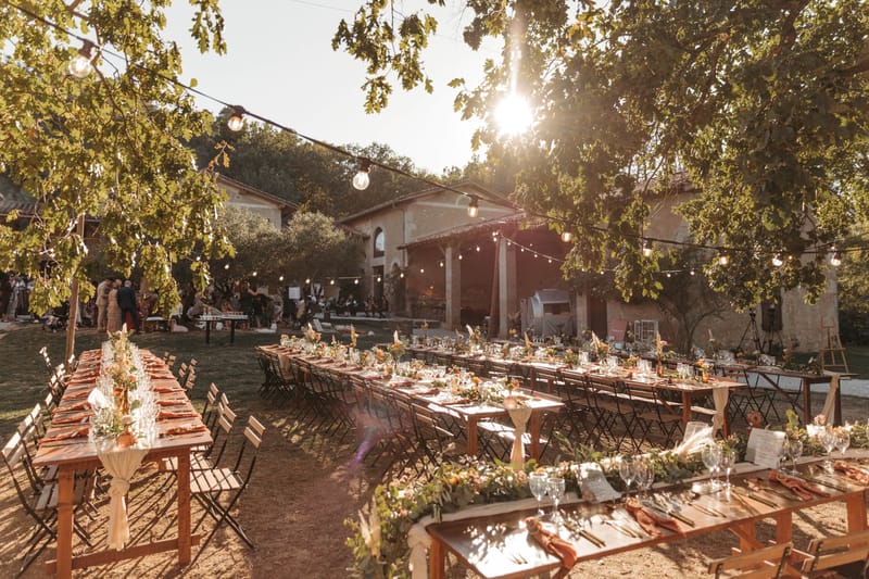 Rustic golden hour reception farm tables with terracotta napkins greenery garlands Edison bulbs and stone mas behind