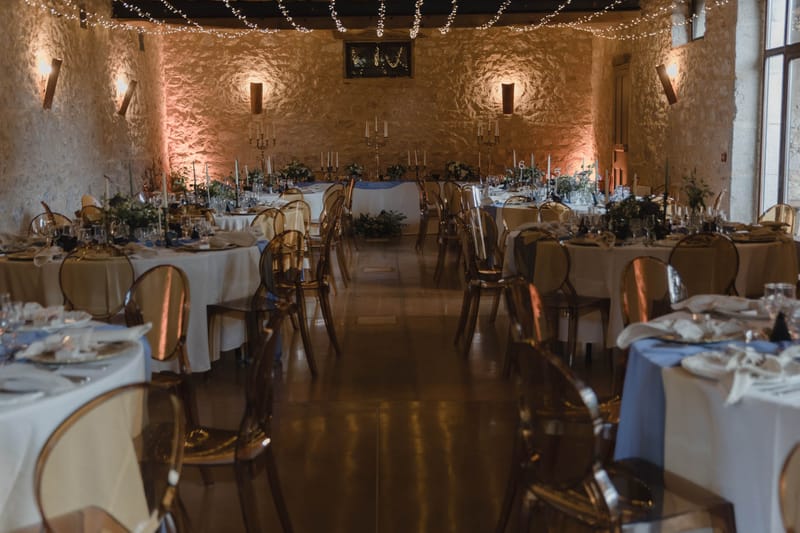 A wide shot of a wedding reception room set up before guests arrive, inside what appears to be a French château or barn venue with exposed limestone walls. The ceiling is draped with fairy lights and warm amber uplighting illuminates the walls, creating a glowing effect. Round guest tables are dressed in white linen with dusty blue table runners, set with glassware, white folded napkins, and greenery-based centerpieces, while transparent amber ghost chairs and gold oval-back chairs are arranged around them. A rectangular head table at the far end features tall silver candelabras with white taper candles, white floral arrangements, and a framed monogram sign on the wall above. The overall decor palette combines white, dusty blue, and gold with a classic-meets-modern styling approach. Potential venue feature image.