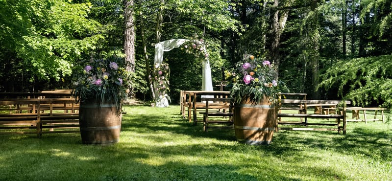 Wine barrel planters with pink peonies and mauve blooms flanking aisle to draped floral arch in woodland