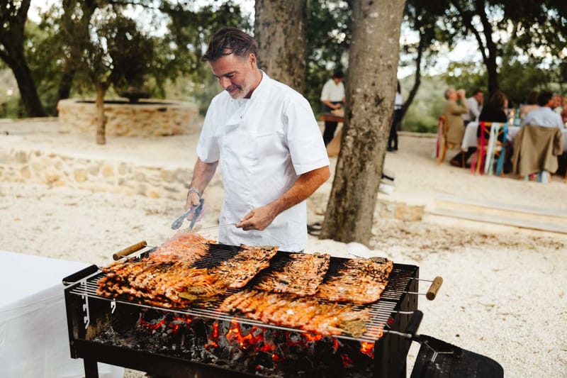 An outdoor wedding reception catering scene at what appears to be a rustic Provençal estate or domaine, with a chef in a white chef's jacket grilling large quantities of whole prawns or langoustines over an open charcoal barbecue with visible glowing embers. The chef is smiling and using metal tongs to tend to the seafood, which covers the entire grill surface. In the background, wedding guests are seated at long tables with natural linen table coverings and colorful chairs — including what appear to be brightly colored bistro-style chairs in yellow, red, and blue — set among mature trees on a gravel and sandy ground. A stone well or water feature is visible to the left. The setting is casual and festive with a relaxed, rustic outdoor dining style. Medium shot with the chef and grill in sharp focus and guests softly blurred in the background.