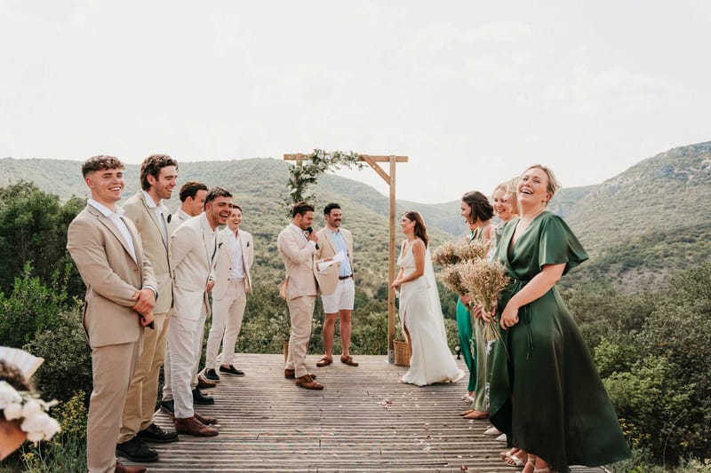 Joyful ceremony on wooden deck with valley backdrop bridesmaids in green satin groomsmen in beige linen under wooden arch