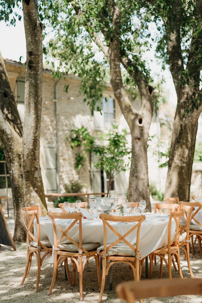 Round reception table with white linen and cross-back chairs set in French country courtyard under mature trees