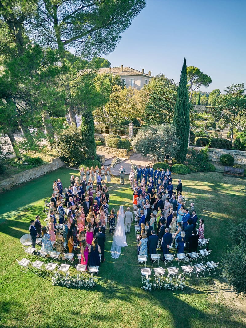Aerial view of outdoor wedding ceremony on a Provencal lawn with bride walking the aisle toward groomsmen and bridesmaids