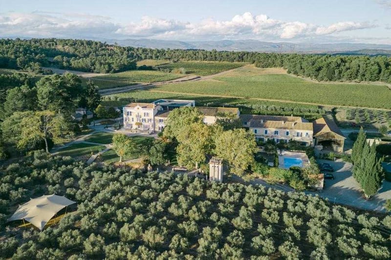 Aerial wide shot of a Provençal mas or domaine property set within an expansive vineyard landscape. The estate comprises a multi-storey stone manor house with cream-painted façade, several adjoining stone outbuildings with terracotta-tiled roofs, and a rectangular swimming pool visible to the right. A large white stretch tent or marquee is pitched in the foreground among rows of olive trees, indicating event or wedding use. The property is accessed via a tree-lined driveway and is surrounded by vineyards extending to the horizon, with forested hills and distant mountains visible in the background. Potential venue feature image.