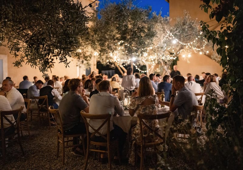 Evening reception under olive trees with festoon lights and 50 guests at round tables on gravel