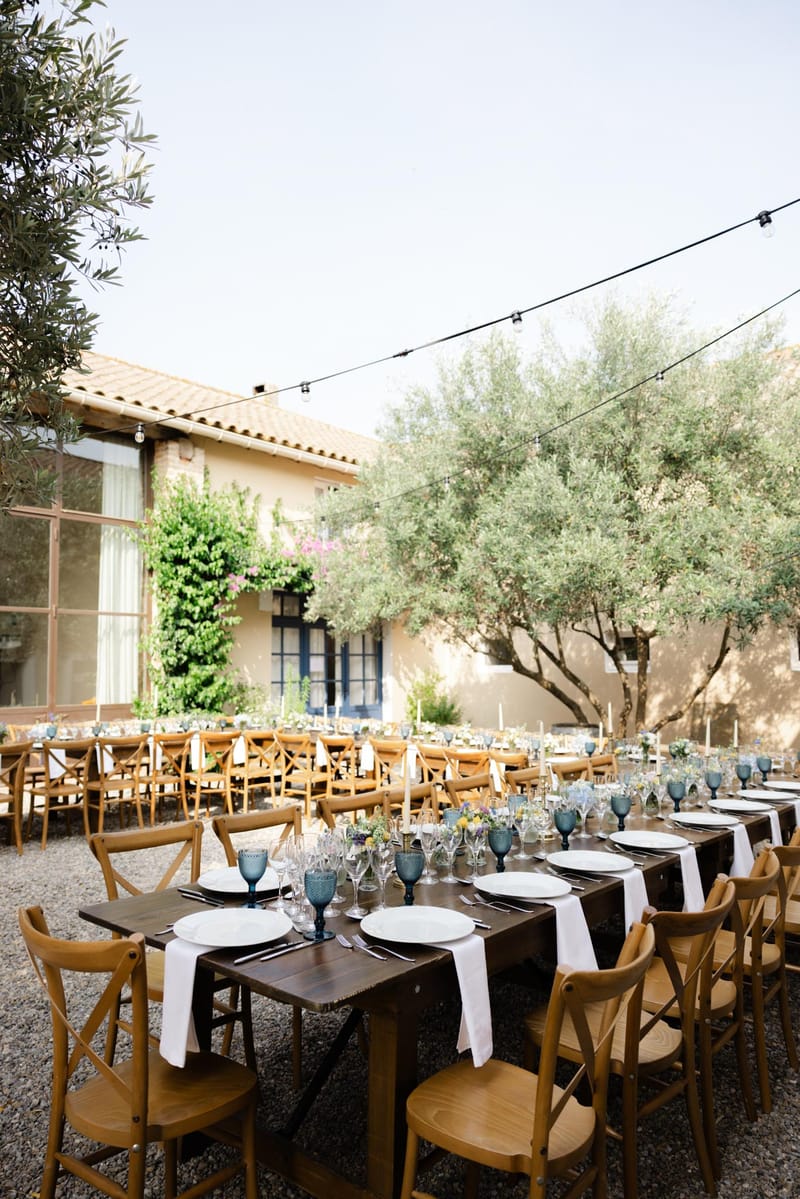Courtyard reception with dark wood farm tables, cross-back chairs, navy glassware, and bistro string lights