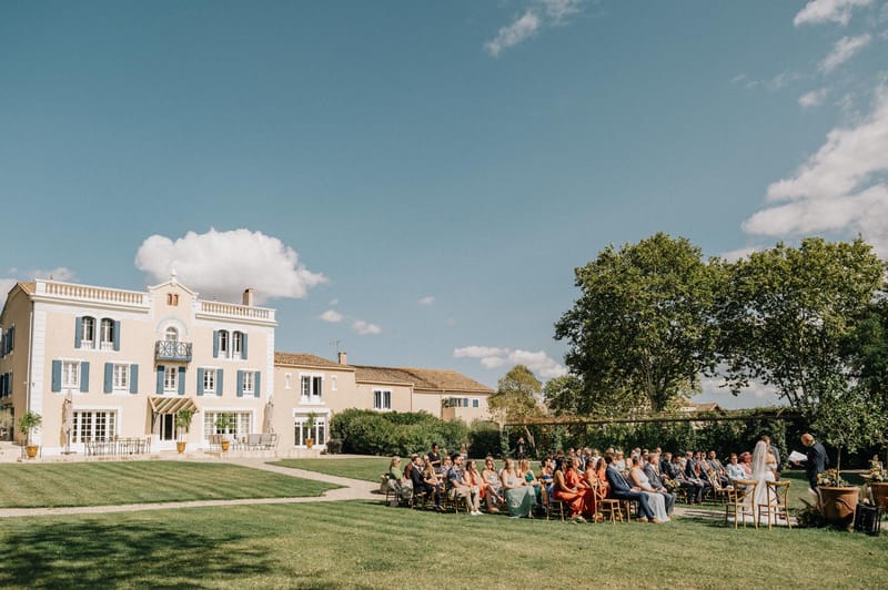 Outdoor wedding ceremony on the lawn of a peach-toned French manor with blue shutters and seated guests