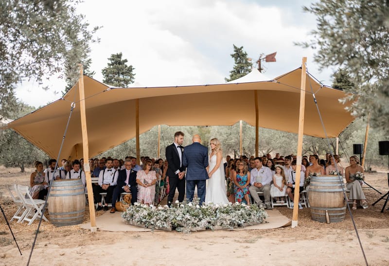 Ceremony under sand stretch tent with white rose platform arrangement oak barrels and olive trees