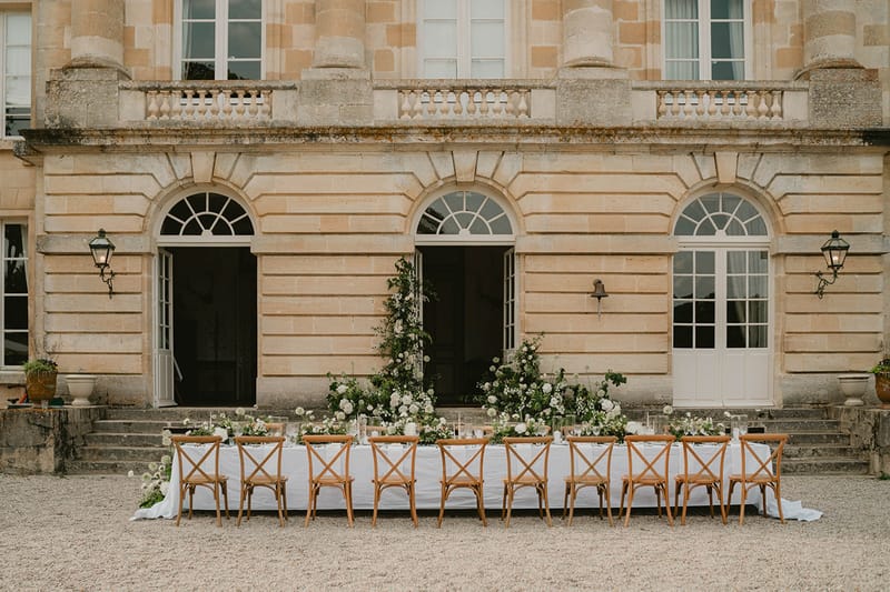 Long table with white rose and allium runner and tall floral installation before chateau with lanterns