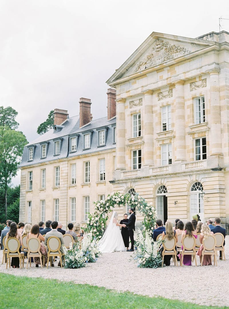 Outdoor wedding ceremony in front of a French chateau with circular floral arch and seated guests on gold chairs