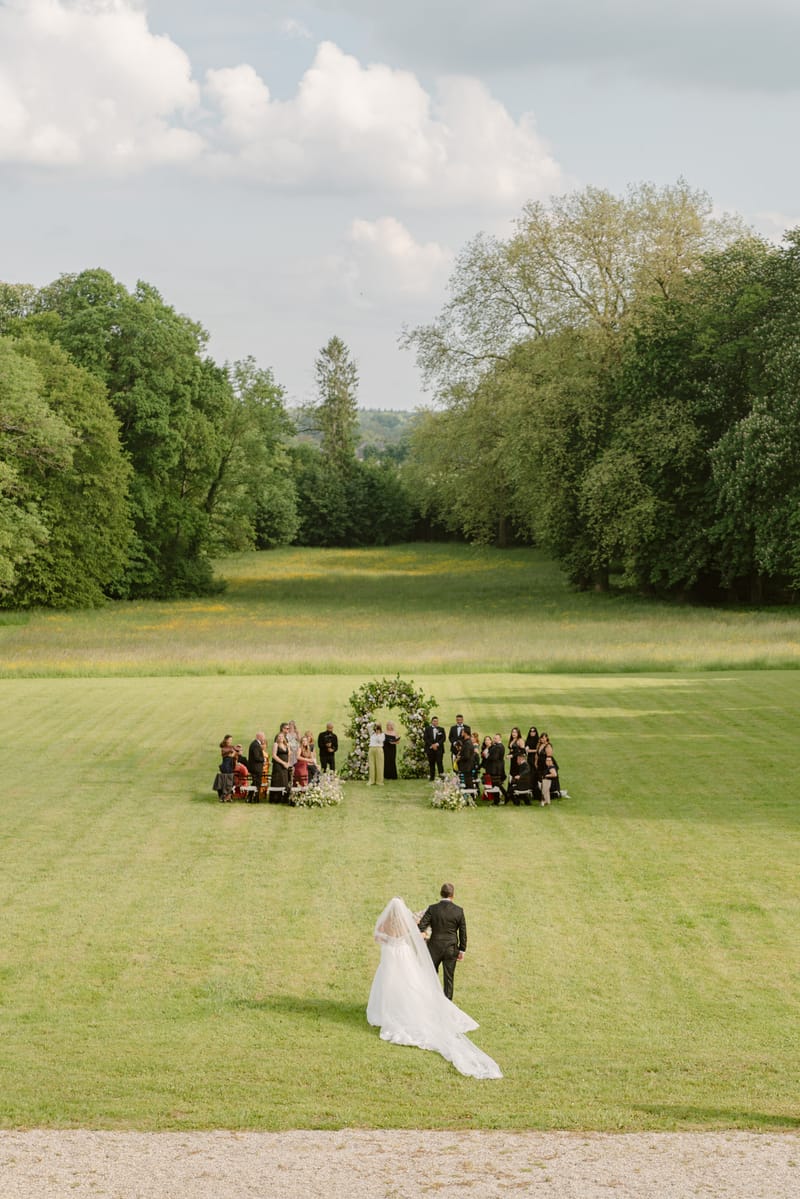 Elevated view of bride walking across lawn toward circular floral arch in white and lavender with seated guests