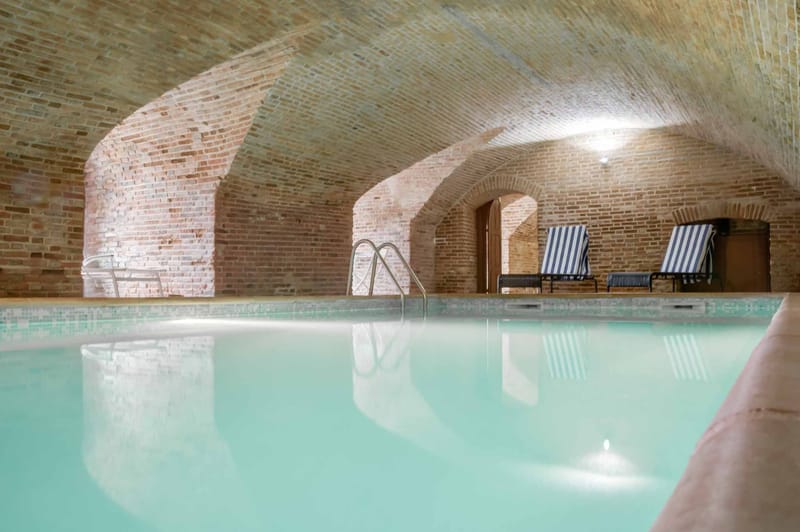 An indoor swimming pool housed within a vaulted brick cellar or undercroft, featuring a series of arched brick ceilings and warm-toned brick walls. The pool water appears a pale turquoise-green, lit from below with a visible underwater light. Two blue-and-white striped sun loungers are positioned at the poolside near an arched doorway in the background, and a metal pool ladder is visible at the far end. The wide-angle shot captures the full length of the pool and the architectural character of the space, with wall-mounted lighting illuminating the brick arches. No people are present. Potential venue feature image.