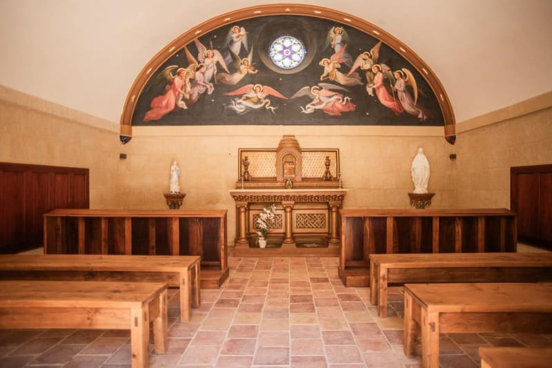 Interior of a small Catholic chapel with gilded altar, angel fresco, rose window, wood pews, and terracotta floor tiles