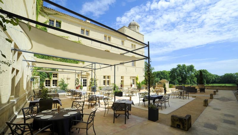 Wide-angle exterior shot of a large French chateau terrace set up for an outdoor reception or dining event. The terrace features multiple round and rectangular tables dressed in dark charcoal and black tablecloths with white plate settings, surrounded by wrought-iron chairs. A large cream-colored sail shade canopy on a black metal frame covers a portion of the terrace. The pale limestone chateau facade, partially covered in climbing ivy, rises several stories and includes a domed turret. Potted topiaries and ornamental plants are arranged around the terrace perimeter, with open parkland and mature trees visible beyond the terrace balustrade. No people are present in the image. Potential venue feature image.
