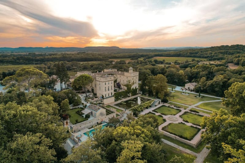 Aerial drone shot of a French château and its surrounding grounds at sunset. The multi-story stone château, built in warm beige limestone, sits at the center of the image, flanked by formal French gardens with geometric lawn panels, gravel pathways, and manicured hedging laid out in a symmetrical pattern. A swimming pool with a tiled surround is visible in the lower left, and additional outbuildings and terraces extend across the estate. The property is surrounded by dense woodland and open countryside, with vineyards and rolling hills visible in the distance. No people or wedding-specific decor are visible in this image. Potential venue feature image.