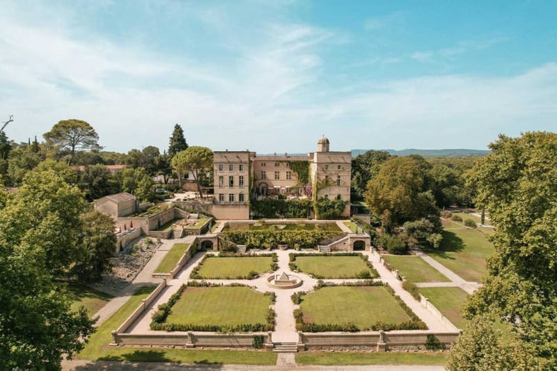 An aerial wide shot of a French château and its formal grounds, with no wedding event activity visible. The multi-storey stone château features a domed tower, terracotta roof tiles, and climbing vines on its facade. In front of the building, a symmetrical formal garden is laid out with four rectangular lawn parterres divided by stone pathways, centered around a circular stone fountain. Stone balustrades and arched brick structures flank the terraced levels connecting the château to the garden. The property is surrounded by mature trees, and a mountain range is visible in the far background. Potential venue feature image.