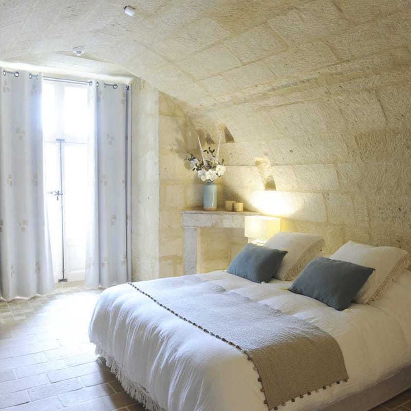 Guest bedroom in French chateau with vaulted stone walls, white linen bed, blue velvet cushions, and stone fireplace
