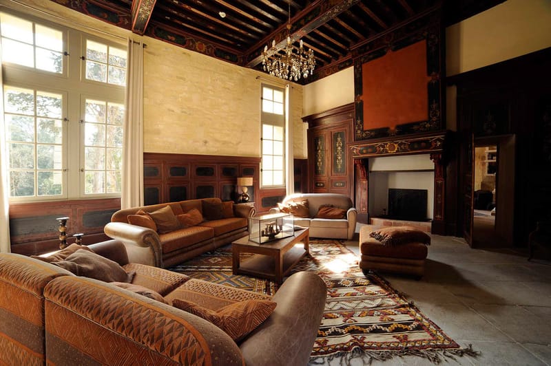 Interior wide shot of a grand salon within a château or historic manor, shown without any wedding guests or couple present. The room features double-height ceilings with dark exposed wooden beams painted with ornate floral and foliate motifs in red, gold, and black. A crystal chandelier hangs centrally from the ceiling. The walls combine exposed limestone with dark wood panelling, also decorated with painted floral patterns, and a large ornate fireplace with an elaborately carved and painted surround anchors the right side of the room. The seating area consists of brown leather and caramel velvet sofas and armchairs arranged around a glass-topped wooden coffee table, all placed on layered Berber-style kilim rugs in cream, red, mustard, and blue geometric patterns. Tall white-framed windows with sheer white curtains line the left wall, allowing warm natural light to stream across the stone-tiled floor. The overall interior style mixes classical French château architecture with warm, eclectic furnishings. Potential venue feature image.