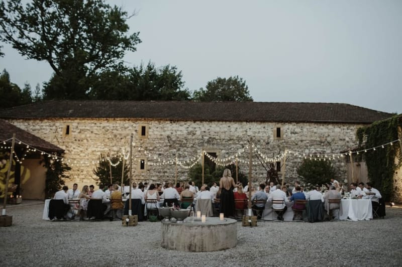 Outdoor wedding reception at dusk in French stone barn courtyard with festoon lights and 50-60 seated guests