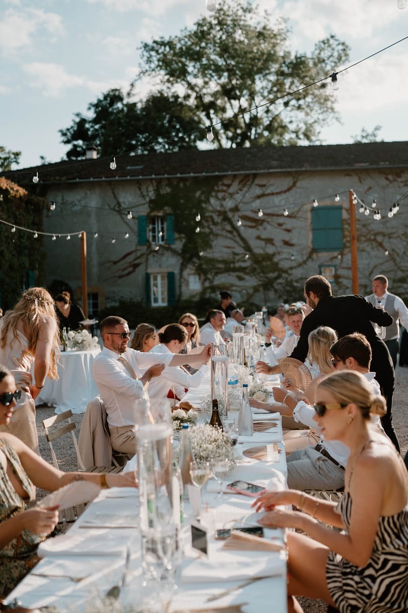 An outdoor wedding reception dinner is underway at a French country property, with approximately 20–25 guests seated along a long banquet-style table covered in white linen. The table is styled with clusters of white baby's breath, tall glass carafes, champagne flutes, and bottles of wine and water as centerpieces. Guests are dressed in a warm, summery palette — many in white, cream, beige, and neutral tones, with one guest in a black-and-white zebra-print dress in the foreground. The setting is a gravel courtyard in front of a stone farmhouse with teal/blue-green shutters and climbing bare vines on the facade. Overhead festoon bulb string lights are strung between wooden posts and across the building, adding warm evening ambiance. The shot is a medium wide-angle taken from one end of the table, capturing the full length of the gathering in natural late-afternoon light.