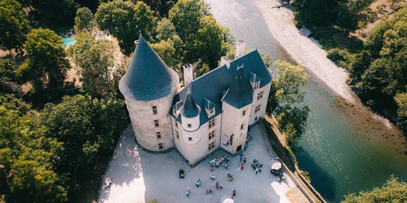 Aerial view of medieval chateau with round tower beside a river, guests gathered on courtyard for cocktail hour