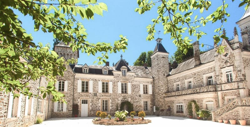 A wide-angle exterior shot of a French château courtyard, framed by tree branches in the foreground. The building is constructed from grey-beige stone with white-shuttered windows, multiple towers with dark slate roofs, decorative stonework detailing, and an ornate exterior staircase on the right wing. The courtyard features a central circular flower bed planted with white and yellow blooms, along with terracotta pots and climbing plants near the entrance archway. No people are visible in the image. Potential venue feature image.