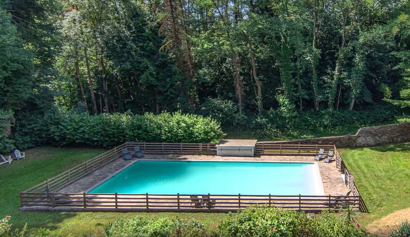 Aerial wide shot of an outdoor rectangular swimming pool with turquoise water, enclosed by a wooden slatted fence. The pool area includes a stone or paved surround with several dark grey sun loungers positioned around the edges, plus a white equipment storage box at the far end. A small bistro table and chairs are visible near the front fence. The pool is set within a private garden with mown lawn areas on either side and a low stone wall to the right. No people are present in the image. Potential venue feature image.