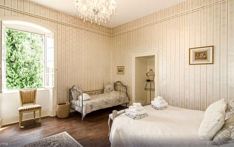 Interior shot of a château bedroom, likely used as a bridal getting-ready suite. The room features cream and taupe vertical striped wallpaper, dark hardwood floors, and a crystal chandelier overhead. Two iron-frame beds are dressed in white and grey paisley linens with white folded towels placed on each, and a larger bed with white lace-edged bedding is visible in the foreground. A vintage gold-frame chair sits near an open arched French window, a wicker basket rests on the floor, a dressmaker's mannequin stands near an interior doorway, and two framed prints hang on the walls. The styling is classic French country with a neutral cream palette throughout. Potential venue feature image.