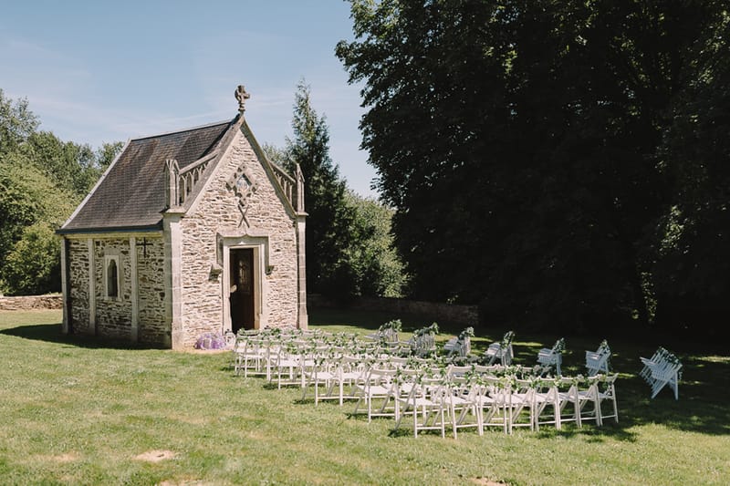 Outdoor ceremony setup with white folding chairs on lawn facing Gothic stone chapel with purple hydrangea accents
