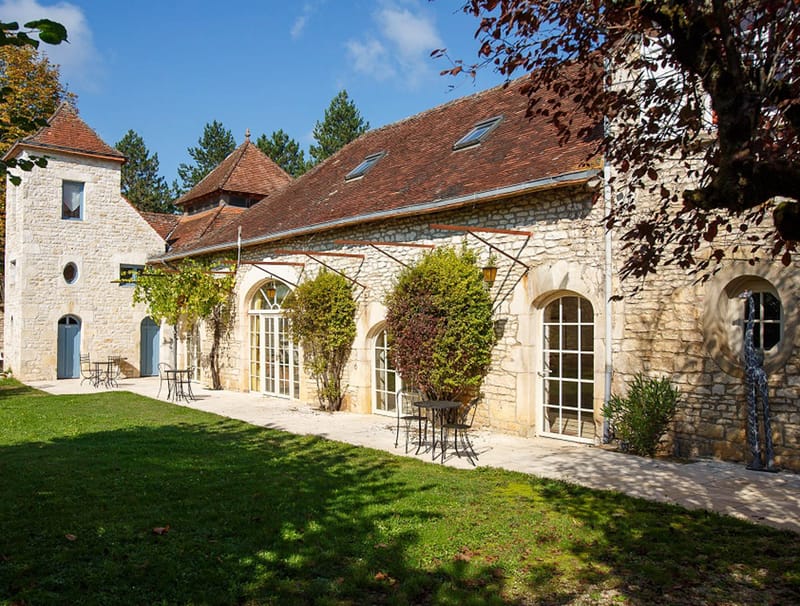 An exterior wide shot of a French stone property featuring a long main building with terracotta-tiled roofing, arched French doors with white-painted frames, and climbing vines trained along the facade. A smaller tower structure with a conical roof stands to the left, fitted with pale blue-grey shuttered doors. A stone-paved terrace runs the length of the building, furnished with small black wrought-iron bistro tables and chairs. No people are visible in the frame. Potential venue feature image.