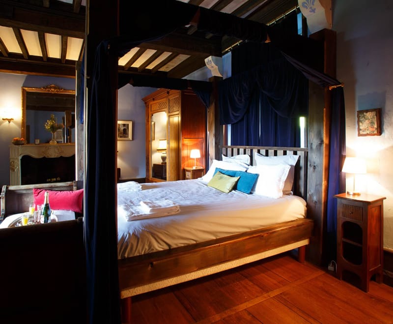 Interior shot of a château guest bedroom styled for a wedding night or honeymoon stay. The room features a large wooden four-poster bed with deep navy blue canopy drapes, dressed in white bed linens with teal, sage green, and teal accent cushions. The room has wide-plank hardwood floors, exposed dark wood ceiling beams with a painted fleur-de-lis motif, and an ornate white stone fireplace visible to the left with a gold-framed mirror above it. A champagne bottle in an ice bucket alongside glasses and a tray is set on a low table near a sofa with a fuchsia pink cushion. Warm ambient lighting comes from bedside table lamps and wall sconces, creating a dark, richly toned atmosphere. A carved wooden armoire and framed artwork complete the classic French château interior styling. Wide interior shot. Potential venue feature image.