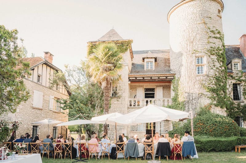 40 guests at long tables under white parasols before ivy-covered tower chateau with deep red centrepieces