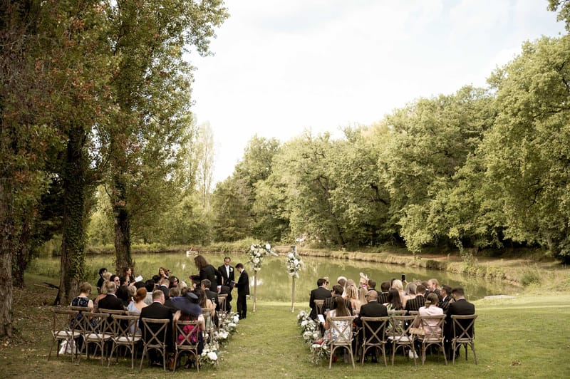 Outdoor riverside wedding ceremony with guests seated on wooden chairs and two grooms at floral arch altar