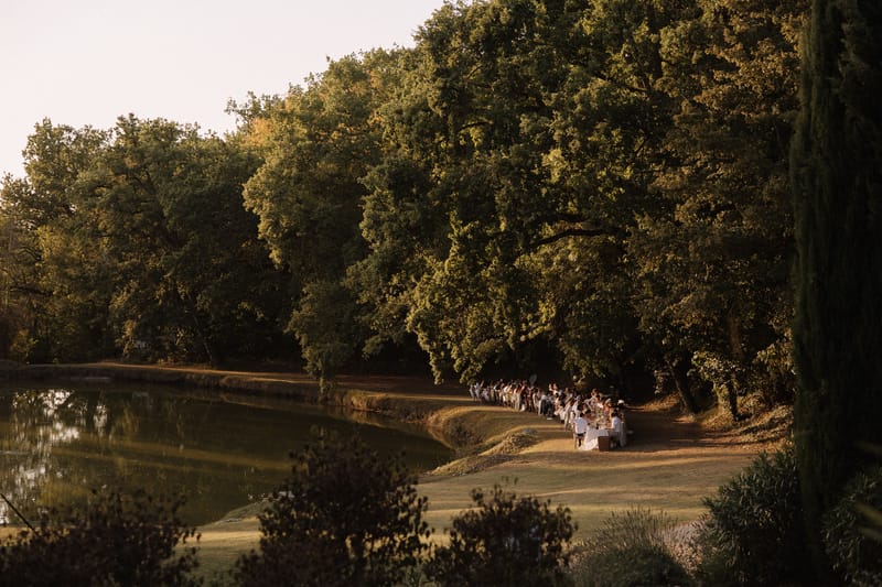 An outdoor wedding reception dinner is taking place along the bank of a calm ornamental lake, photographed from a wide elevated vantage point at golden hour. A very long single banquet table — seating what appears to be 60 or more guests — stretches in a curved line along the water's edge, with white table linens and floral centerpieces visible at the near end. Guests are seated along both sides of the table, dressed predominantly in white and light-colored attire. The setting is a formal French estate or château grounds, with large mature oak trees forming a dense canopy backdrop behind the table. The warm late-afternoon light bathes the scene in golden tones, contrasting with the deep shadow of the tree line. The composition is a wide landscape shot that emphasizes the scale of the tablescape and its relationship to the lakeside grounds. Potential venue feature image.