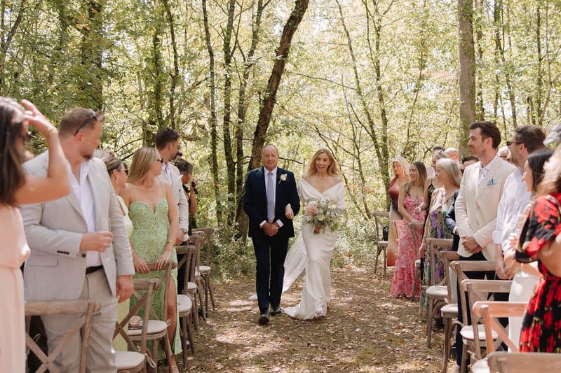 Bride in draped cape gown walked down woodland aisle with cross-back chairs by father in navy suit