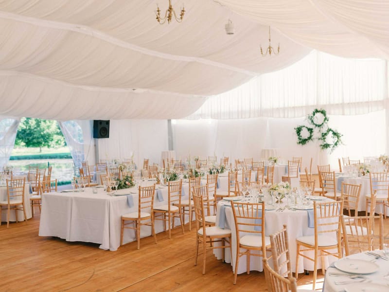 White marquee with oak parquet floor, greenery wreath backdrop, and round tables with blue napkins