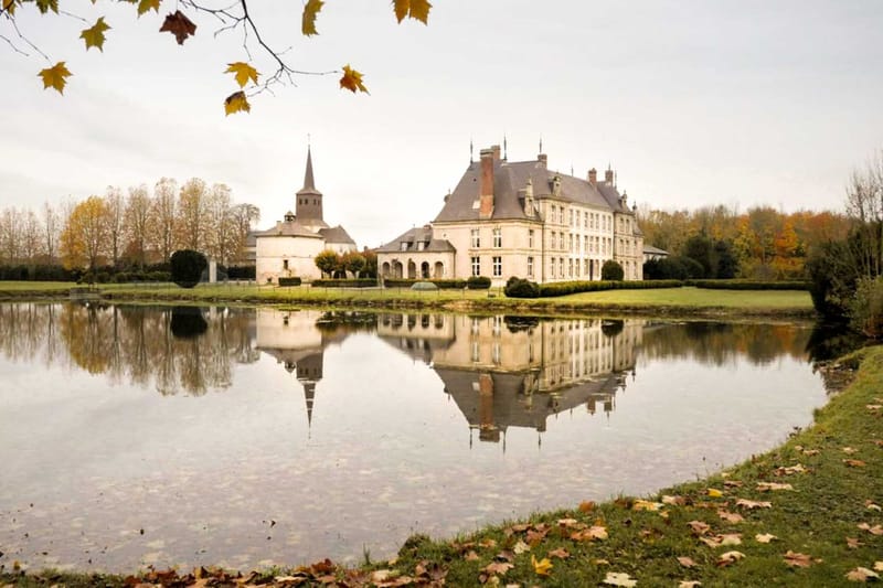 A wide exterior shot of a French château photographed from across a still ornamental lake, with the building and its slate mansard roofs reflected clearly in the water. The château is a multi-storey pale limestone structure with dormer windows, decorative chimney stacks, and pointed roof finials, accompanied by a round tower and a church steeple to its left. No people are visible in the image; this is a grounds and architecture shot taken in autumn, as indicated by fallen leaves in the foreground and yellow-orange foliage surrounding the estate. Potential venue feature image.