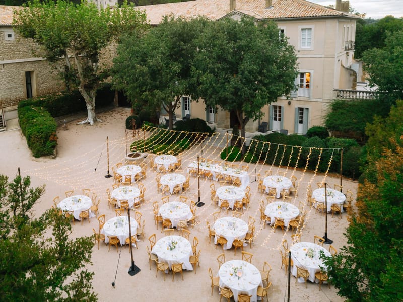 Aerial dusk view of round reception tables under fairy light canopy in Provencal chateau courtyard