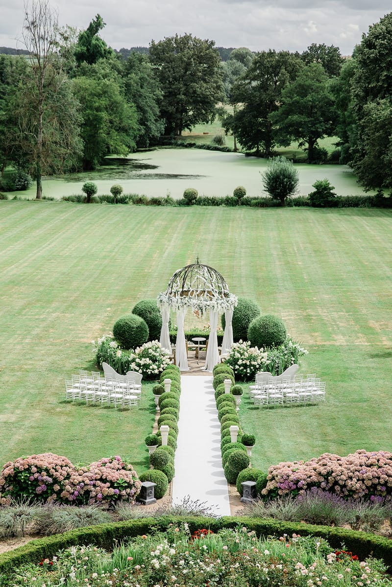 Aerial view of outdoor ceremony setup with white draped gazebo, floral arrangements, and chiavari chairs by ornamental lake