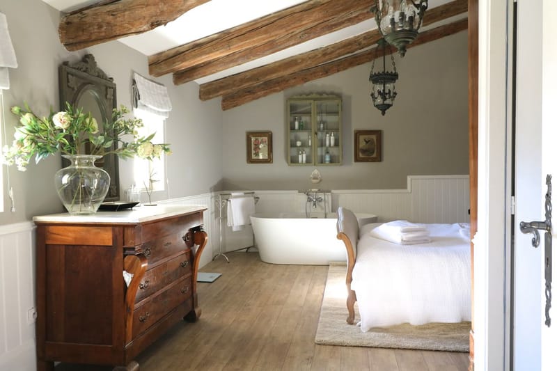 This is not a wedding photograph but rather an interior room shot, likely used as a venue feature image. The room combines bedroom and bathroom spaces in a French country style, featuring exposed rustic wooden ceiling beams, wide-plank hardwood flooring, and white tongue-and-groove wainscoting. A freestanding white soaking tub with floor-mounted chrome fixtures sits in the background, flanked by a wrought-iron towel rail with white towels. In the foreground, an antique walnut chest of drawers with a white marble top holds a clear glass vase filled with cream peonies and green foliage. Additional furnishings include a carved grey-painted mirror, a Louis XV-style upholstered bench in grey linen, a patinated green glass-front cabinet, a hanging lantern-style chandelier, and two small gilt-framed artworks on the wall. The overall palette is warm grey, white, and natural wood tones in a classic French provincial style. Wide shot taken from the doorway. Potential venue feature image.