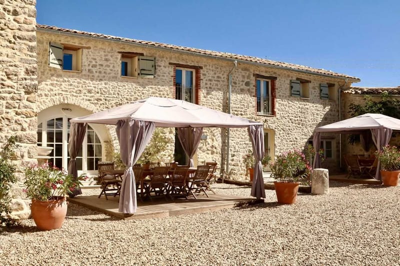 Stone farmhouse courtyard with gazebo canopies sheltering wooden dining table and oleander plants in terracotta pots