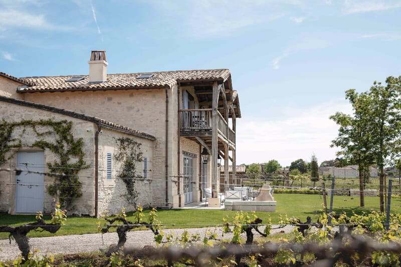 A wide exterior shot of a French country property featuring a two-story stone building with terracotta roof tiles, a chimney, and pale grey shutters. The facade is partially covered with climbing vines, and a rustic open timber-framed gallery structure runs along the side of the building, featuring a first-floor balcony with weathered wooden beams. At ground level, white outdoor lounge furniture and a stone decorative feature are visible on a paved terrace area beside tall French doors. Grapevines on wire trellises are visible in the foreground, indicating a working vineyard setting. The overall architectural style is rustic Provençal farmhouse. Potential venue feature image.
