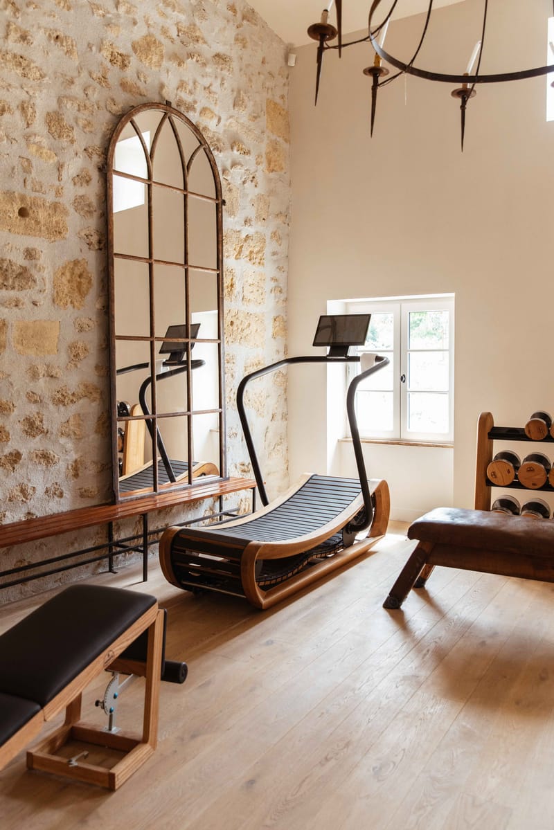 This image does not depict a wedding photograph. It shows the interior of a fitness or wellness room, likely within a French château or manor property. The room features a curved wooden-framed treadmill, a black upholstered weight bench, a wooden dumbbell rack, and a large arched mirror with a dark iron frame leaning against an exposed limestone stone wall. The space has wide-plank light wood flooring, a wrought iron chandelier overhead, and a white-framed French window letting in natural light. The overall aesthetic blends historic architectural features with high-end fitness equipment. Potential venue feature image.
