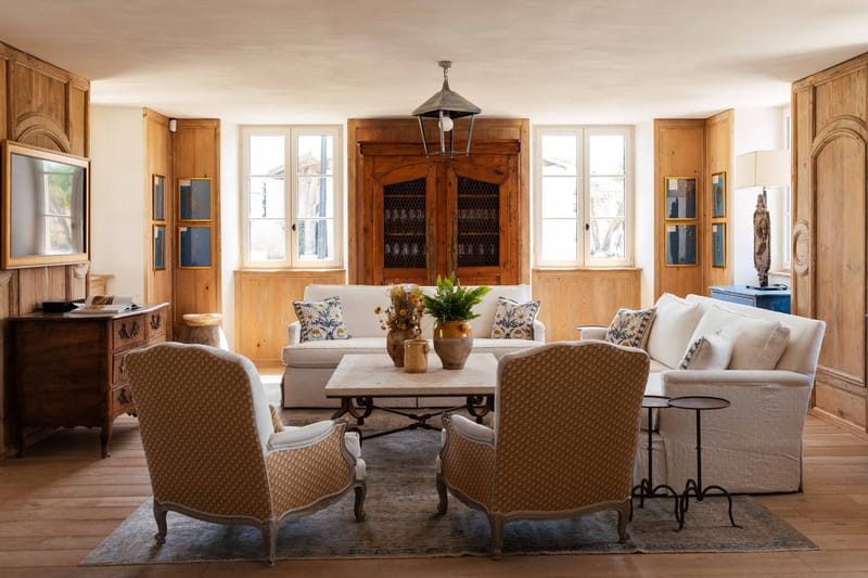 French country salon with honey wood paneling, linen sofas, Louis XV armchairs, and antique pine armoire